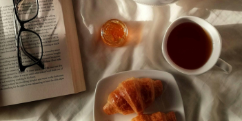 A coffee, crossiant, book with glasses and honey on a bed.